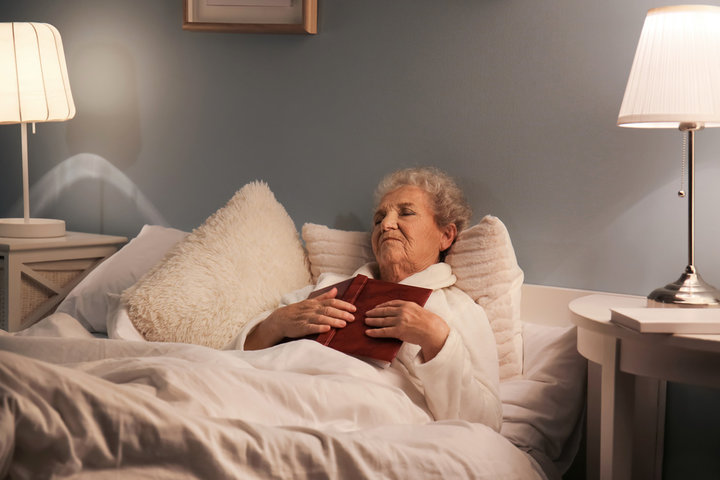 older woman reading a book and smiling and falling asleep in her bed in a care home