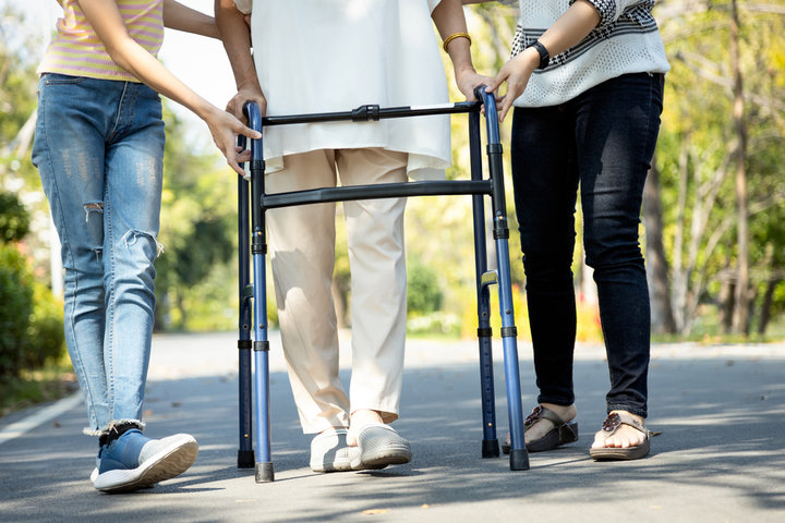 older person using a walking aid in the park with two carers helping support them
